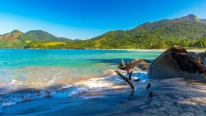 Uma vista panorâmica de uma praia tropical. A areia escura, quase preta, encontra o mar turquesa e translúcido, que forma pequenas ondas brancas. À direita, grandes rochas escuras se elevam da areia, e no centro, pedaços de madeira seca e envelhecida estão presos na areia. Ao fundo, uma exuberante floresta tropical cobre montanhas que se estendem até onde a vista alcança, sob um céu azul claro e ensolarado.