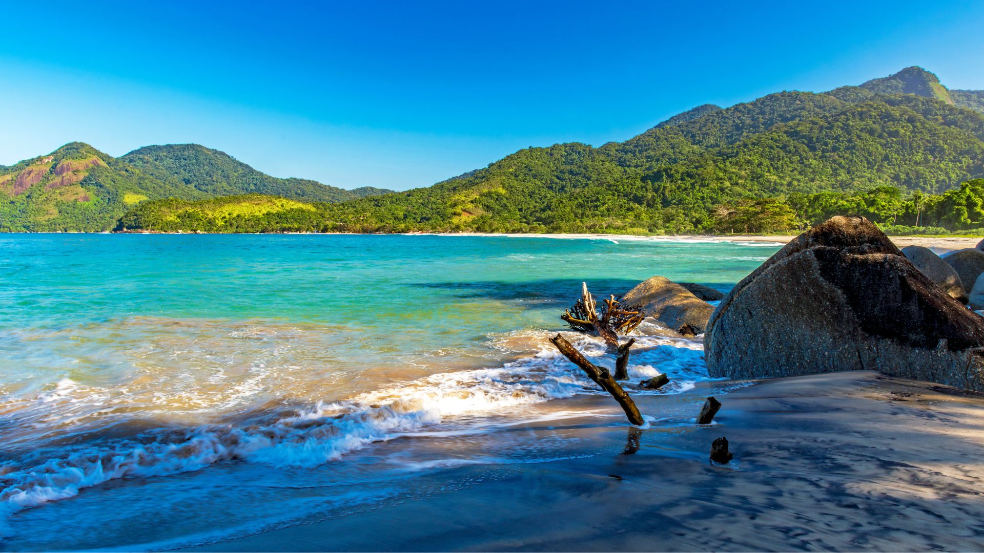 Uma vista panorâmica de uma praia tropical. A areia escura, quase preta, encontra o mar turquesa e translúcido, que forma pequenas ondas brancas. À direita, grandes rochas escuras se elevam da areia, e no centro, pedaços de madeira seca e envelhecida estão presos na areia. Ao fundo, uma exuberante floresta tropical cobre montanhas que se estendem até onde a vista alcança, sob um céu azul claro e ensolarado.