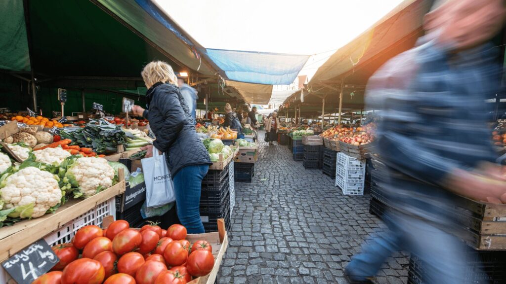 Pessoas fazendo compras de frutas e verduras em um mercado ao ar livre.