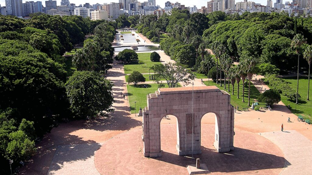 Parque com um grande arco no meio, cercado por árvores e bancos.