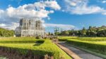 O Jardim Botânico de Curitiba se estende sob um céu azul pontilhado de nuvens brancas e fofas. No centro, a estufa principal, com sua estrutura metálica e vidro, brilha sob o sol. Seus três domos curvos, que lembram palácios de cristal, abrigam uma variedade de plantas. Em primeiro plano, um caminho de pedra ladeado por arbustos bem cuidados convida a um passeio tranquilo. Ao fundo, árvores frondosas completam a paisagem, oferecendo sombra e frescor aos visitantes.
