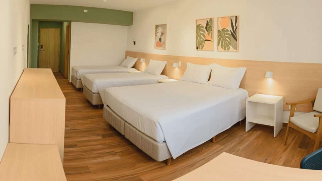 Um quarto de hotel bem iluminado com três camas bem feitas, piso de madeira, uma pequena mesa e arte tropical nas paredes em um ambiente moderno.
