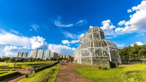 Fotografia ampla do Jardim Botânico de Curitiba em um dia ensolarado. A imagem mostra o jardim em primeiro plano, com um caminho de pedra claro levando em direção à estufa de vidro, o principal destaque do local. A estufa, com sua estrutura de metal branco e curvas ornamentadas, se destaca contra o céu azul com nuvens brancas. Pessoas caminham pelo jardim e pela trilha, e ao fundo, prédios altos e a vegetação exuberante do parque podem ser vistos.