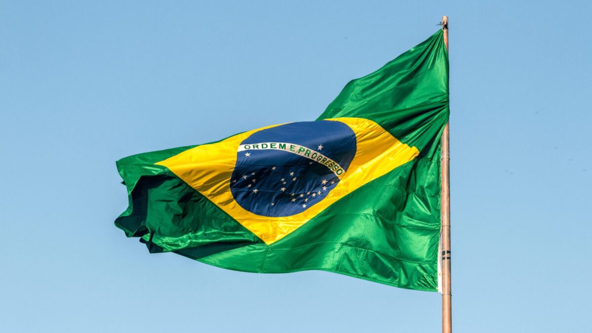 Uma bandeira brasileira de cores vivas ondeia contra um céu azul claro. A bandeira é predominantemente verde, com um grande losango amarelo no centro. Dentro do losango há um círculo azul contendo 27 estrelas brancas e uma faixa branca que diz "ORDEM E PROGRESSO".