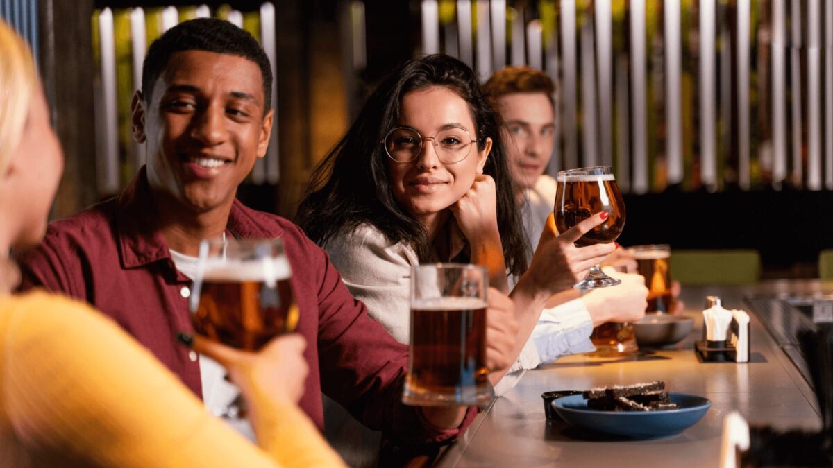 Um grupo de amigos em um bar, brindando com grandes copos de cerveja, desfrutando de bebidas e conversas, com petiscos na mesa.