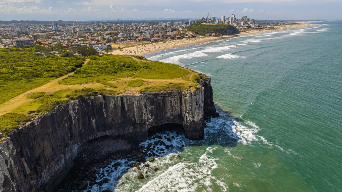 A imagem mostra um penhasco com vista para o oceano. O penhasco é rochoso e tem alguma vegetação no topo. O oceano é azul e tem algumas ondas. No fundo, há uma cidade com prédios e uma praia. A imagem foi tirada de um ângulo alto, olhando para o penhasco e o oceano.