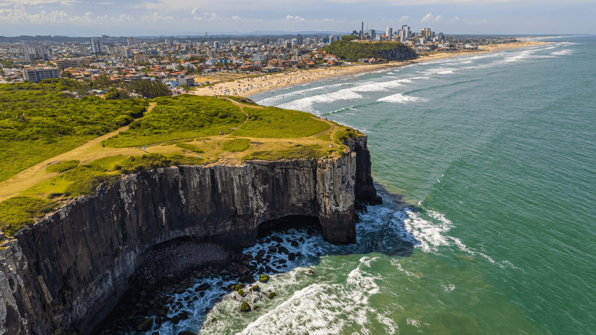 A imagem mostra um penhasco com vista para o oceano. O penhasco é rochoso e tem alguma vegetação no topo. O oceano é azul e tem algumas ondas. No fundo, há uma cidade com prédios e uma praia. A imagem foi tirada de um ângulo alto, olhando para o penhasco e o oceano.
