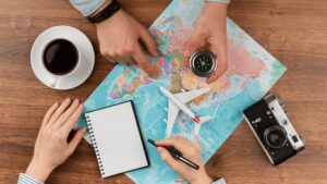 Mãos apontando para um mapa-múndi com uma câmera, café, um caderno, um modelo de avião e uma bússola, sugerindo planejamento de viagem.