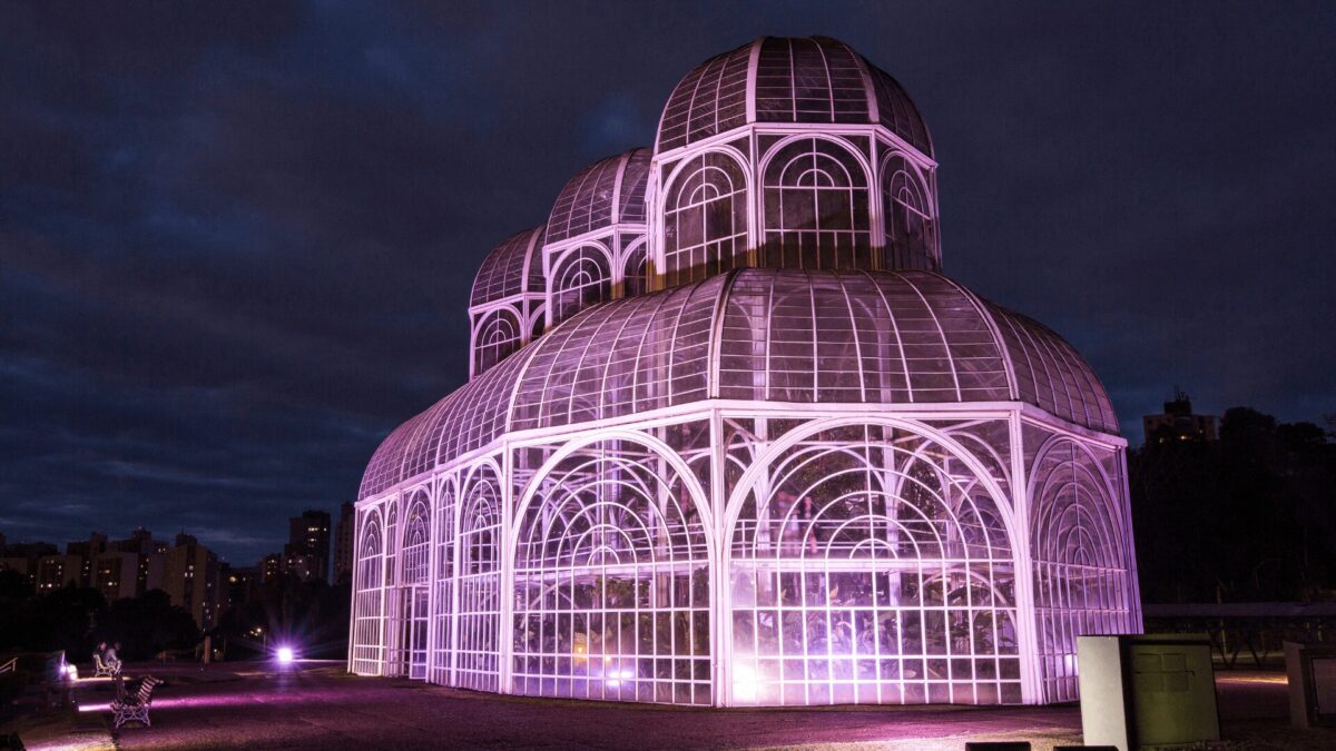 Uma estufa de vidro lindamente iluminada brilha em rosa contra um céu crepuscular, cercada por prédios da cidade ao fundo.