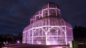 Uma estufa de vidro lindamente iluminada brilha em rosa contra um céu crepuscular, cercada por prédios da cidade ao fundo.