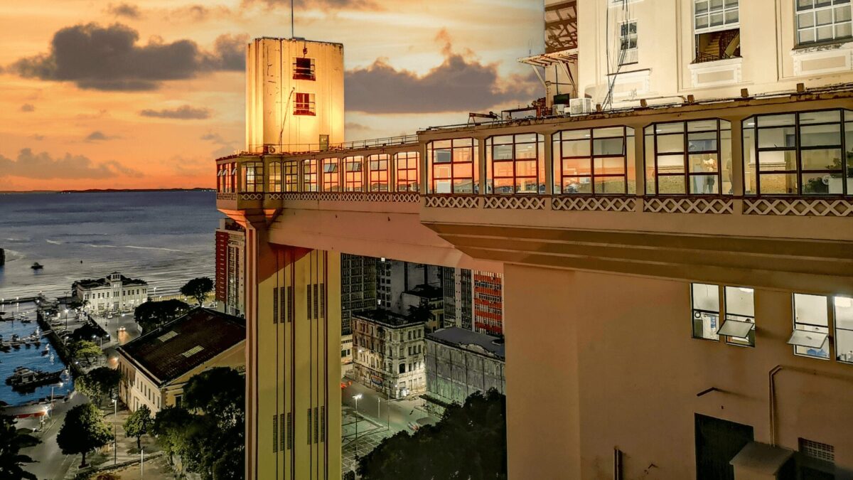 Uma vista do pôr do sol do icônico elevador de Salvador, mostrando sua estrutura e o porto próximo, com cores quentes refletindo na água.