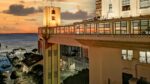 Uma vista do pôr do sol do icônico elevador de Salvador, mostrando sua estrutura e o porto próximo, com cores quentes refletindo na água.