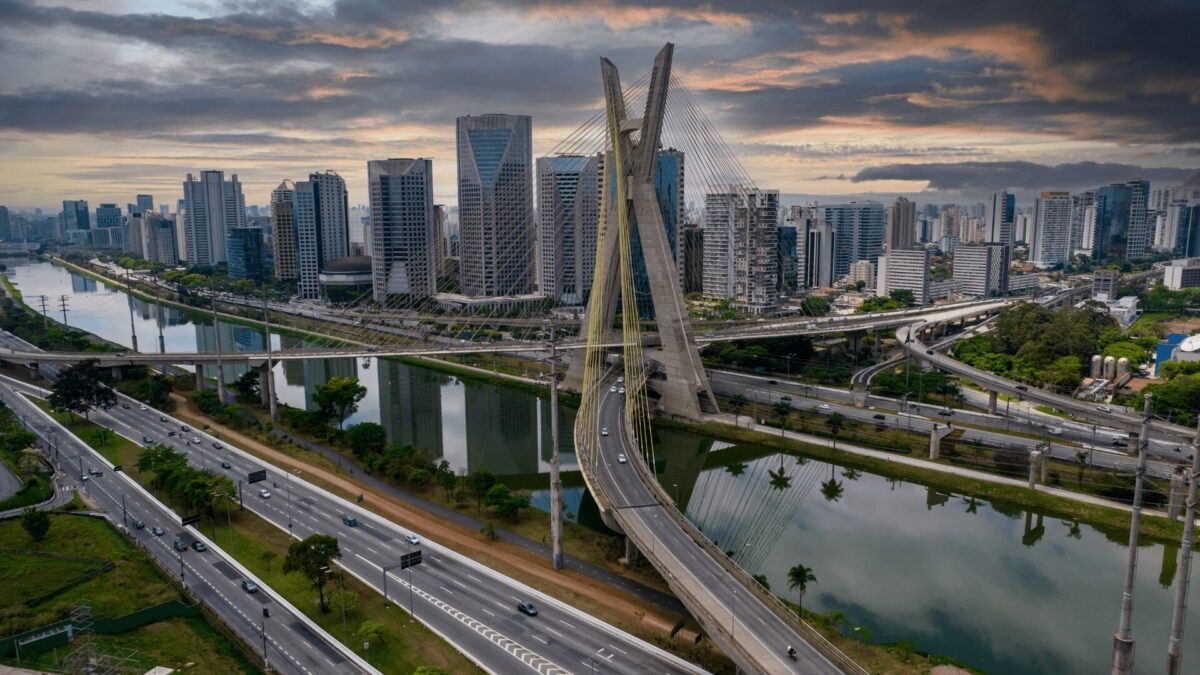 Vista aérea da icônica ponte e do horizonte de São Paulo, refletidos no rio sob um céu dramático. Estradas movimentadas com tráfego abaixo.