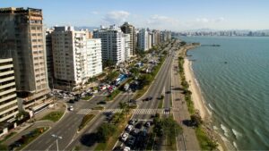 A imagem mostra uma vista aérea diurna da Avenida Beira-Mar Norte em Florianópolis, Santa Catarina, Brasil. A avenida, com múltiplas pistas e tráfego intenso de veículos, corre paralela à costa, que se estende à direita da imagem. À esquerda da avenida, erguem-se altos edifícios residenciais e comerciais, alguns em construção, característicos de uma área urbana densa. Vegetação e árvores podem ser vistas entre os prédios e ao longo da avenida. No centro, a avenida em si é o elemento dominante, com carros, ônibus e outros veículos movendo-se em ambas as direções. Faixas de pedestres são visíveis em cruzamentos. À direita da avenida, uma faixa de areia clara forma a praia, seguida pelas águas da Baía Norte, que apresentam uma tonalidade verde-azulada. Ao longo da orla, há uma ciclovia e uma área de calçadão com pessoas caminhando e árvores espaçadas. Ao longe, no horizonte, é possível avistar a silhueta de mais áreas urbanas e morros. O céu está azul e claro, com boa visibilidade, indicando um dia ensolarado. A perspectiva da foto é de cima e ligeiramente angulada, proporcionando uma visão ampla da paisagem urbana e costeira.