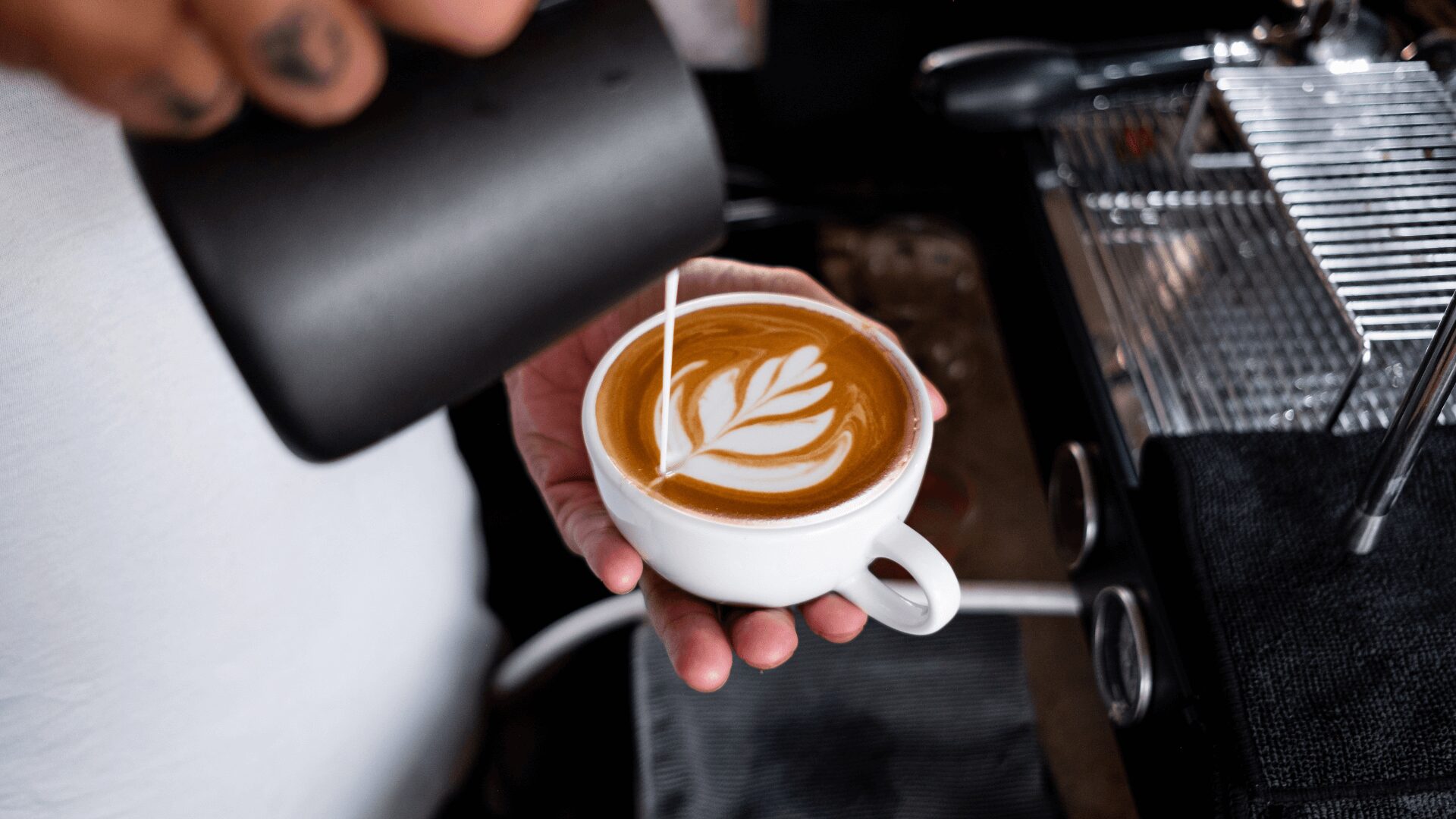 Um barista despeja leite vaporizado em uma xícara de café, criando uma complexa arte latte em uma superfície marrom quente.