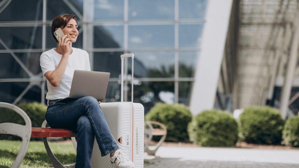Uma pessoa sentada em um banco com um laptop e uma mala, falando ao telefone, tendo como pano de fundo arquitetura moderna e muito verde.