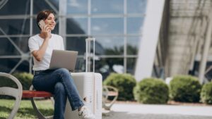 Uma pessoa sentada em um banco com um laptop e uma mala, falando ao telefone, tendo como pano de fundo arquitetura moderna e muito verde.