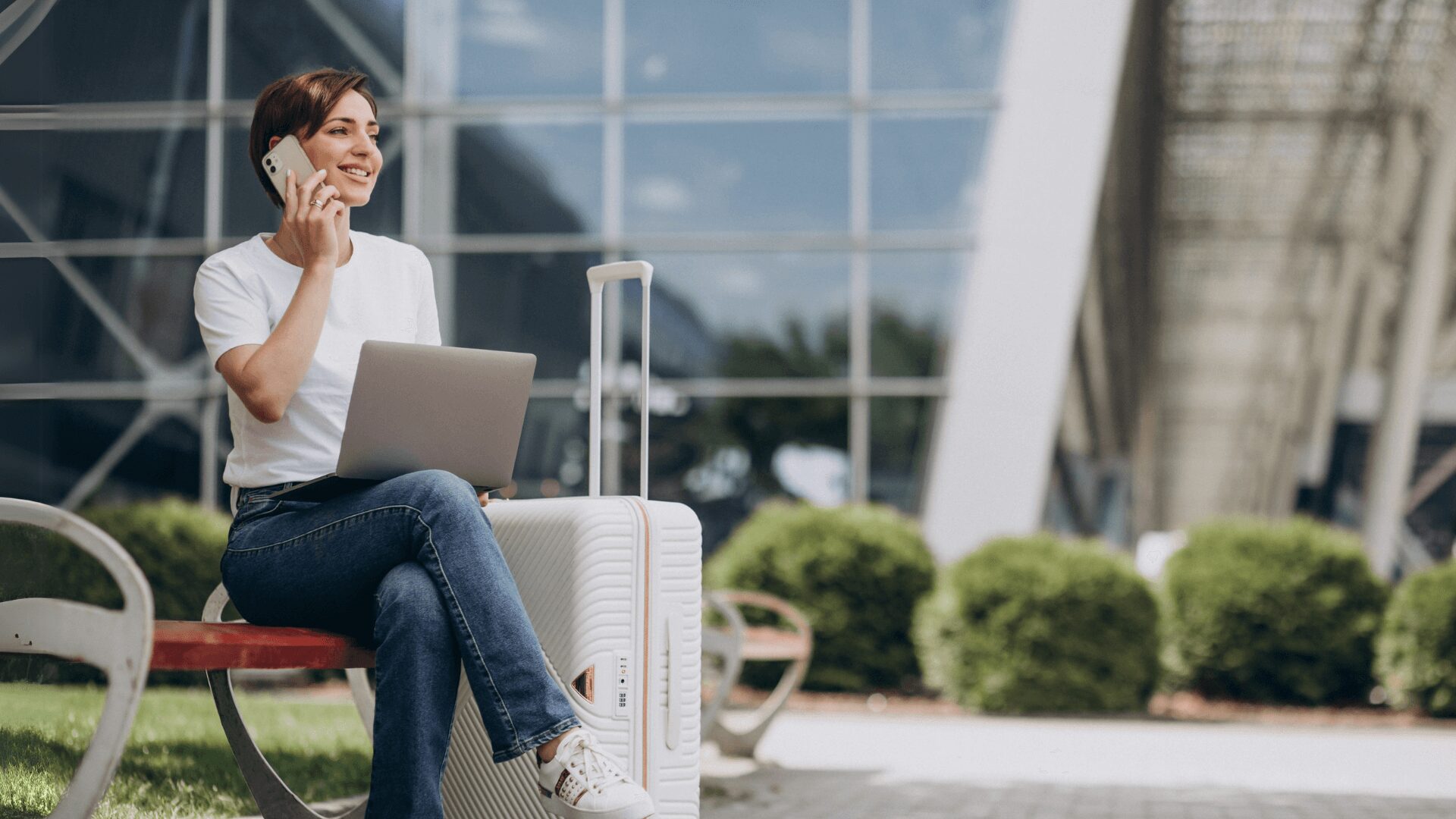 Uma pessoa sentada em um banco com um laptop e uma mala, falando ao telefone, tendo como pano de fundo arquitetura moderna e muito verde.
