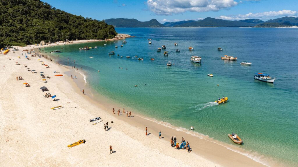 Um cenário de praia vibrante com pessoas tomando sol, nadando e grupos se misturando, cercados por águas azuis cristalinas e barcos ao longe.