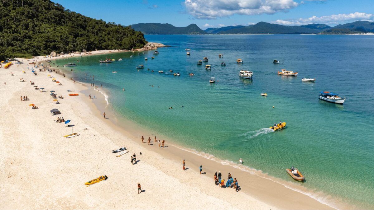Um cenário de praia vibrante com pessoas tomando sol, nadando e grupos se misturando, cercados por águas azuis cristalinas e barcos ao longe.