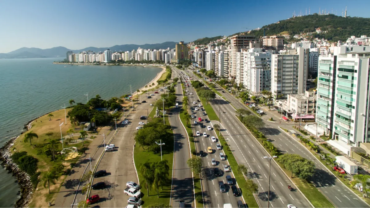 Vista aérea de uma estrada costeira ladeada por prédios altos, carros, parques verdes e o mar sob um céu azul claro.
