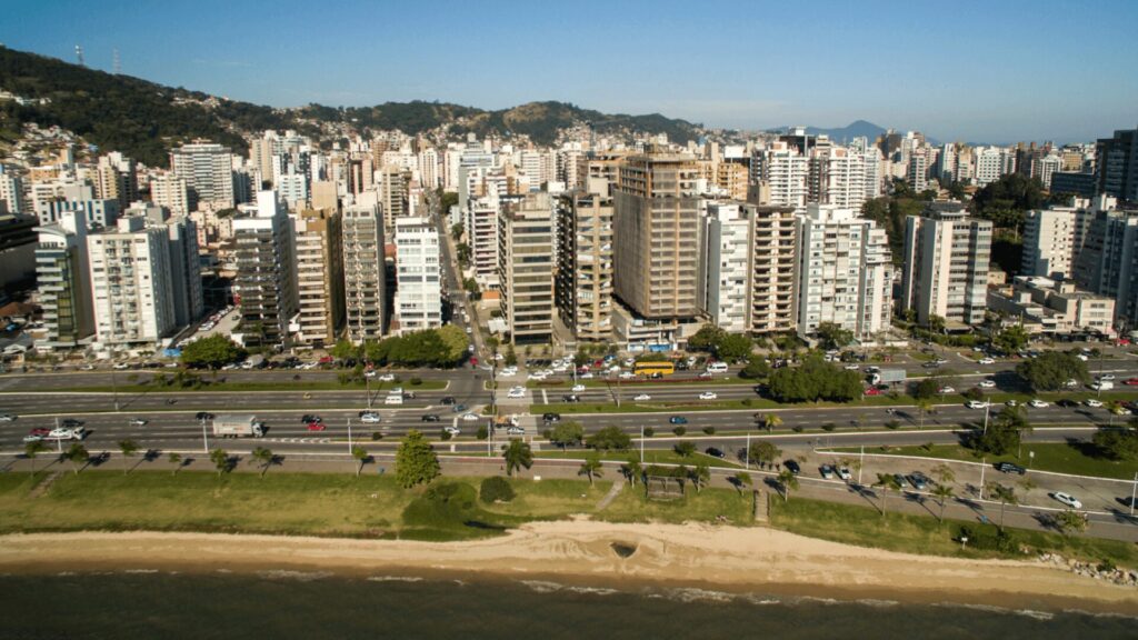 Vista aérea de uma cidade costeira com edifícios modernos, uma estrada movimentada ladeada por árvores e uma praia de areia à beira da água.