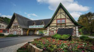 Uma foto da entrada para a cidade de Campos do Jordão, com um grande portal de estilo europeu, que se assemelha a casas com telhados pontiagudos e detalhes em madeira e cores claras. Um letreiro verde com "Campos do Jordão" em branco está pendurado acima da estrada que passa por baixo do portal. Em primeiro plano, há um canteiro de flores com flores vermelhas e brancas, e um pequeno vaso de madeira preta com grama verde. O céu é azul com algumas nuvens.