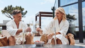 Um casal de roupões brancos brinda com taças de champanhe sentado ao ar livre, cercado por uma decoração moderna e uma paisagem serena.