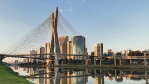 A imagem exibe a Ponte Octávio Frias de Oliveira, também conhecida como Ponte Estaiada, em São Paulo, Brasil, sob um céu claro e ensolarado. A ponte se destaca com seus dois mastros curvos que se encontram no topo, de onde partem inúmeros cabos de aço que sustentam o tabuleiro. Sua estrutura é refletida nitidamente na superfície calma da água abaixo. Ao fundo e à direita da ponte, há um aglomerado de arranha-céus modernos e de vidro que também se refletem na água, criando um panorama urbano elegante. Mais à direita, um complexo de viadutos e ruas elevadas adiciona camadas à paisagem urbana. A margem esquerda do rio é coberta por grama verde, contrastando com o ambiente construído. A iluminação dourada do sol da tarde sugere um fim de tarde ou começo de manhã, conferindo à cena uma atmosfera serena e majestosa.