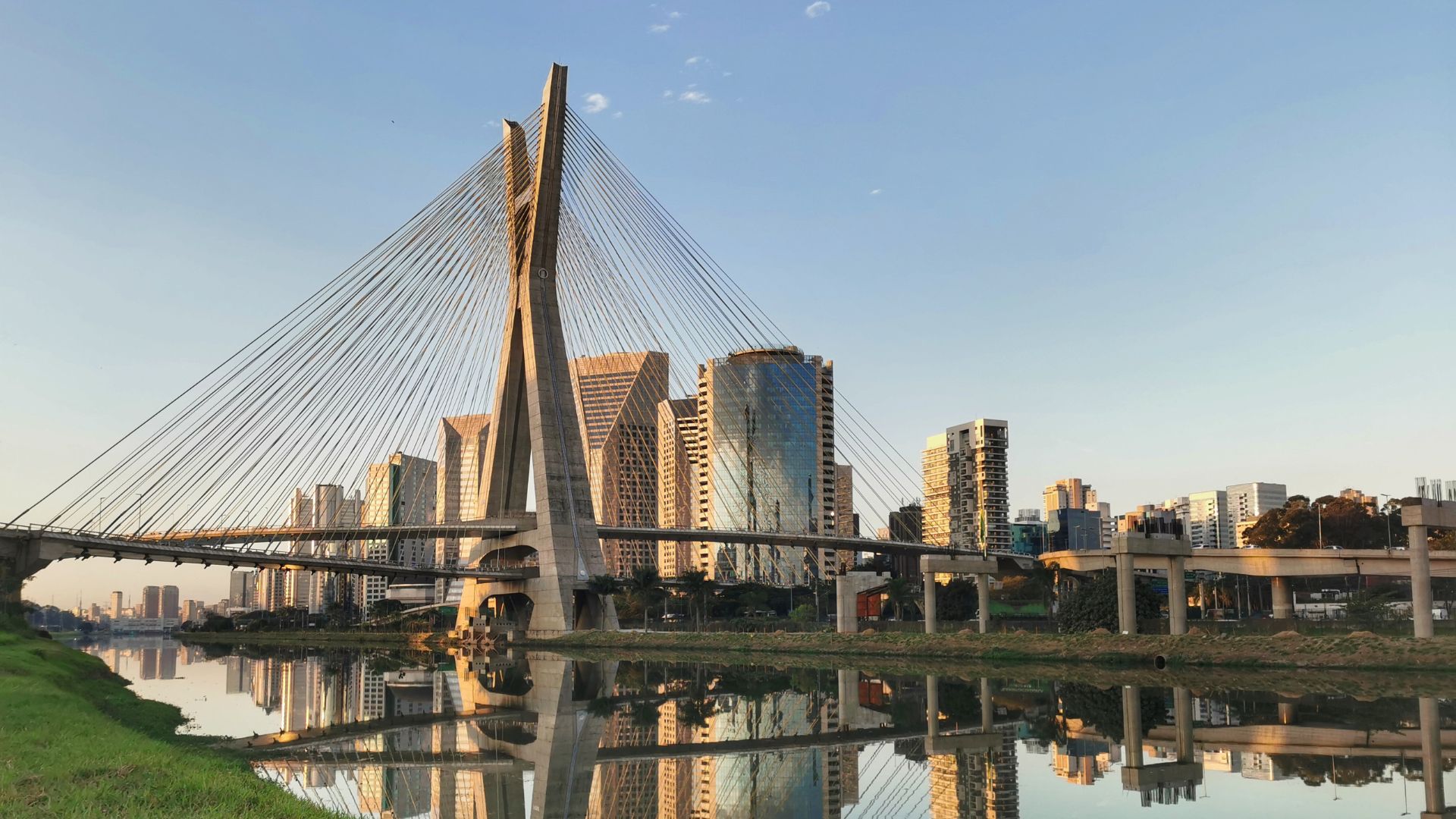 A imagem exibe a Ponte Octávio Frias de Oliveira, também conhecida como Ponte Estaiada, em São Paulo, Brasil, sob um céu claro e ensolarado. A ponte se destaca com seus dois mastros curvos que se encontram no topo, de onde partem inúmeros cabos de aço que sustentam o tabuleiro. Sua estrutura é refletida nitidamente na superfície calma da água abaixo. Ao fundo e à direita da ponte, há um aglomerado de arranha-céus modernos e de vidro que também se refletem na água, criando um panorama urbano elegante. Mais à direita, um complexo de viadutos e ruas elevadas adiciona camadas à paisagem urbana. A margem esquerda do rio é coberta por grama verde, contrastando com o ambiente construído. A iluminação dourada do sol da tarde sugere um fim de tarde ou começo de manhã, conferindo à cena uma atmosfera serena e majestosa.