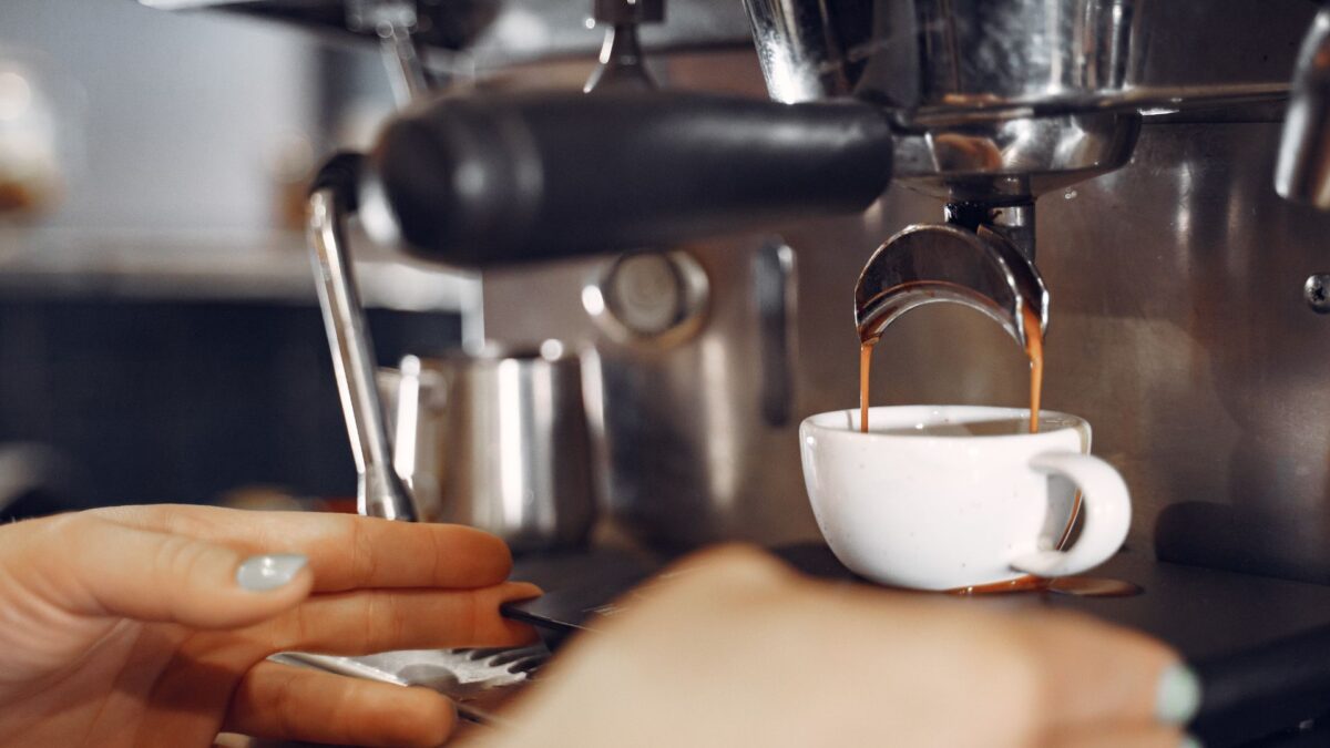 Uma foto em close-up e com foco seletivo captura o momento em que uma máquina de café profissional prepara um expresso. No centro da imagem, um fluxo duplo de café marrom-escuro e cremoso escorre do bico de um porta-filtro de metal para dentro de uma pequena xícara de cerâmica branca. A xícara está posicionada sobre a grade da máquina. Em primeiro plano, a mão de uma pessoa com unhas pintadas de um tom cinza-azulado claro repousa suavemente perto da base da xícara. Partes de outra mão também são visíveis, mas desfocadas. O fundo da imagem mostra o corpo robusto e metálico da máquina de café expresso, com seus botões e alavancas, e uma pequena jarra de metal para vaporizar leite ao lado, tudo suavemente desfocado para dar destaque à extração do café. A iluminação é suave e destaca o brilho do metal e a riqueza da cor do café, criando uma atmosfera convidativa e artesanal.