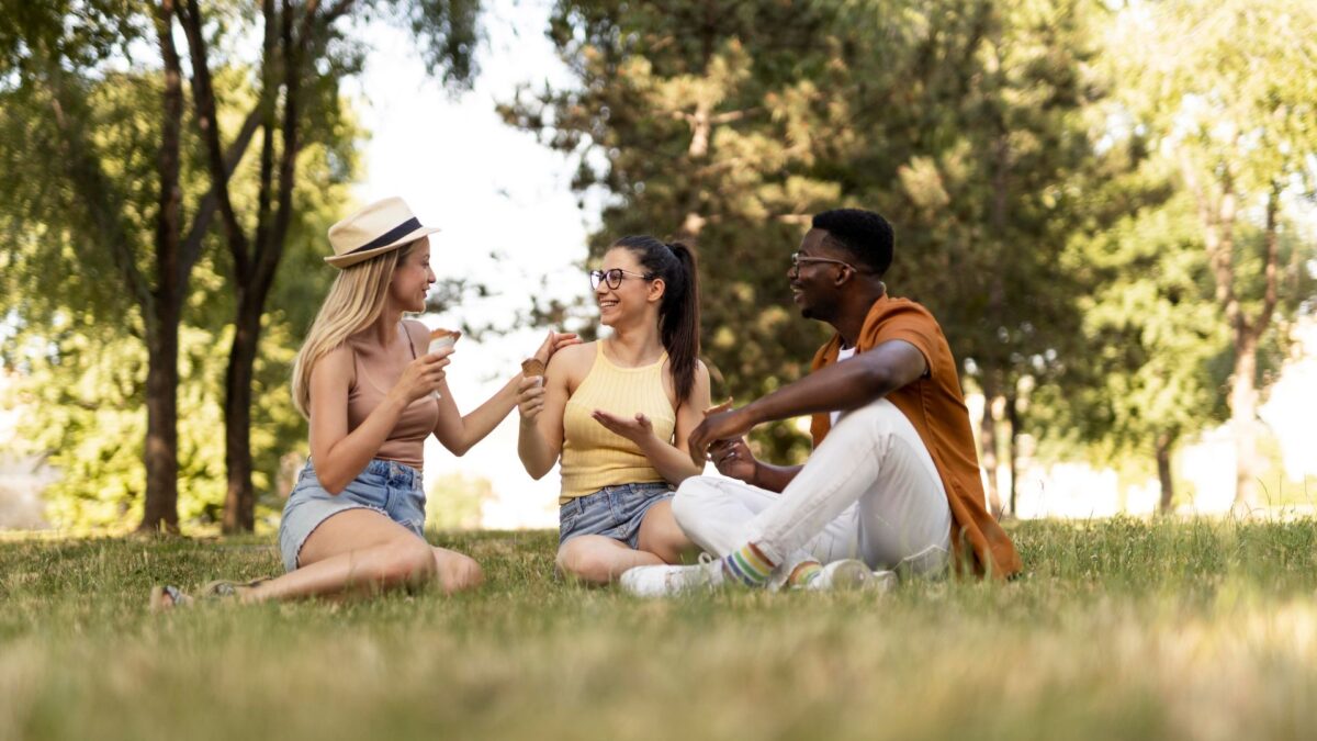 A imagem mostra três jovens, dois deles parecendo ser mulheres e um homem, sentados na grama em um parque sob a sombra de árvores frondosas.