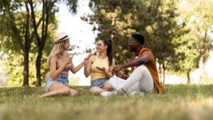 A imagem mostra três jovens, dois deles parecendo ser mulheres e um homem, sentados na grama em um parque sob a sombra de árvores frondosas.
