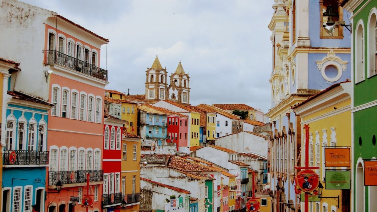 Uma vista panorâmica e colorida do Pelourinho, em Salvador, Bahia. A imagem exibe uma rua inclinada com edifícios históricos de fachadas em tons vibrantes como rosa, azul-turquesa, amarelo, laranja, verde e branco. No topo da colina, destacam-se as duas torres brancas de uma igreja barroca, com telhados em terracota. O céu está nublado, e alguns detalhes arquitetônicos como balcões e janelas estão visíveis, com bandeiras ou estandartes coloridos pendurados nas ruas estreitas.