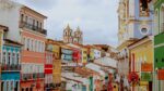 Uma vista panorâmica e colorida do Pelourinho, em Salvador, Bahia. A imagem exibe uma rua inclinada com edifícios históricos de fachadas em tons vibrantes como rosa, azul-turquesa, amarelo, laranja, verde e branco. No topo da colina, destacam-se as duas torres brancas de uma igreja barroca, com telhados em terracota. O céu está nublado, e alguns detalhes arquitetônicos como balcões e janelas estão visíveis, com bandeiras ou estandartes coloridos pendurados nas ruas estreitas.