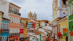 Uma vista panorâmica e colorida do Pelourinho, em Salvador, Bahia. A imagem exibe uma rua inclinada com edifícios históricos de fachadas em tons vibrantes como rosa, azul-turquesa, amarelo, laranja, verde e branco. No topo da colina, destacam-se as duas torres brancas de uma igreja barroca, com telhados em terracota. O céu está nublado, e alguns detalhes arquitetônicos como balcões e janelas estão visíveis, com bandeiras ou estandartes coloridos pendurados nas ruas estreitas.