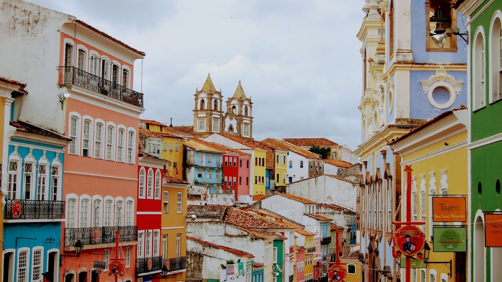 Uma vista panorâmica e colorida do Pelourinho, em Salvador, Bahia. A imagem exibe uma rua inclinada com edifícios históricos de fachadas em tons vibrantes como rosa, azul-turquesa, amarelo, laranja, verde e branco. No topo da colina, destacam-se as duas torres brancas de uma igreja barroca, com telhados em terracota. O céu está nublado, e alguns detalhes arquitetônicos como balcões e janelas estão visíveis, com bandeiras ou estandartes coloridos pendurados nas ruas estreitas.