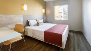 Um quarto de hotel moderno e funcional do Go Inn Curitiba. A imagem mostra uma cama de casal com lençóis brancos e uma manta vermelha dobrada aos pés, um travesseiro enrolado marrom claro no centro da cama. Há duas luminárias de parede acesas acima da cabeceira cinza clara. À esquerda, uma mesa de trabalho com tampo claro e uma cadeira de design moderno em tons de bege. Ao fundo, uma grande janela permite a entrada de luz natural e revela parte da arquitetura de edifícios urbanos. O piso é de madeira clara.