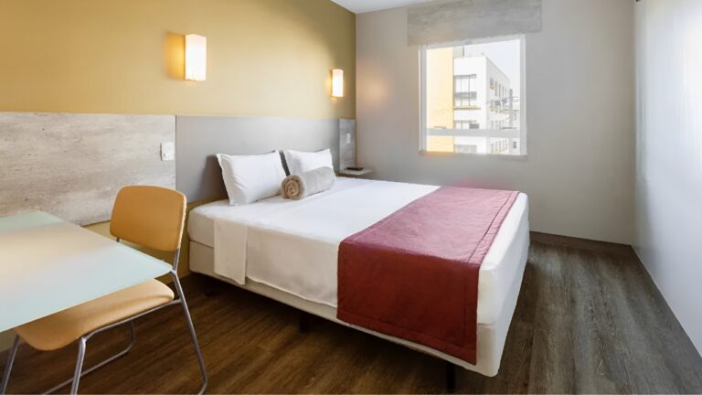 Um quarto de hotel moderno e funcional do Go Inn Curitiba. A imagem mostra uma cama de casal com lençóis brancos e uma manta vermelha dobrada aos pés, um travesseiro enrolado marrom claro no centro da cama. Há duas luminárias de parede acesas acima da cabeceira cinza clara. À esquerda, uma mesa de trabalho com tampo claro e uma cadeira de design moderno em tons de bege. Ao fundo, uma grande janela permite a entrada de luz natural e revela parte da arquitetura de edifícios urbanos. O piso é de madeira clara.