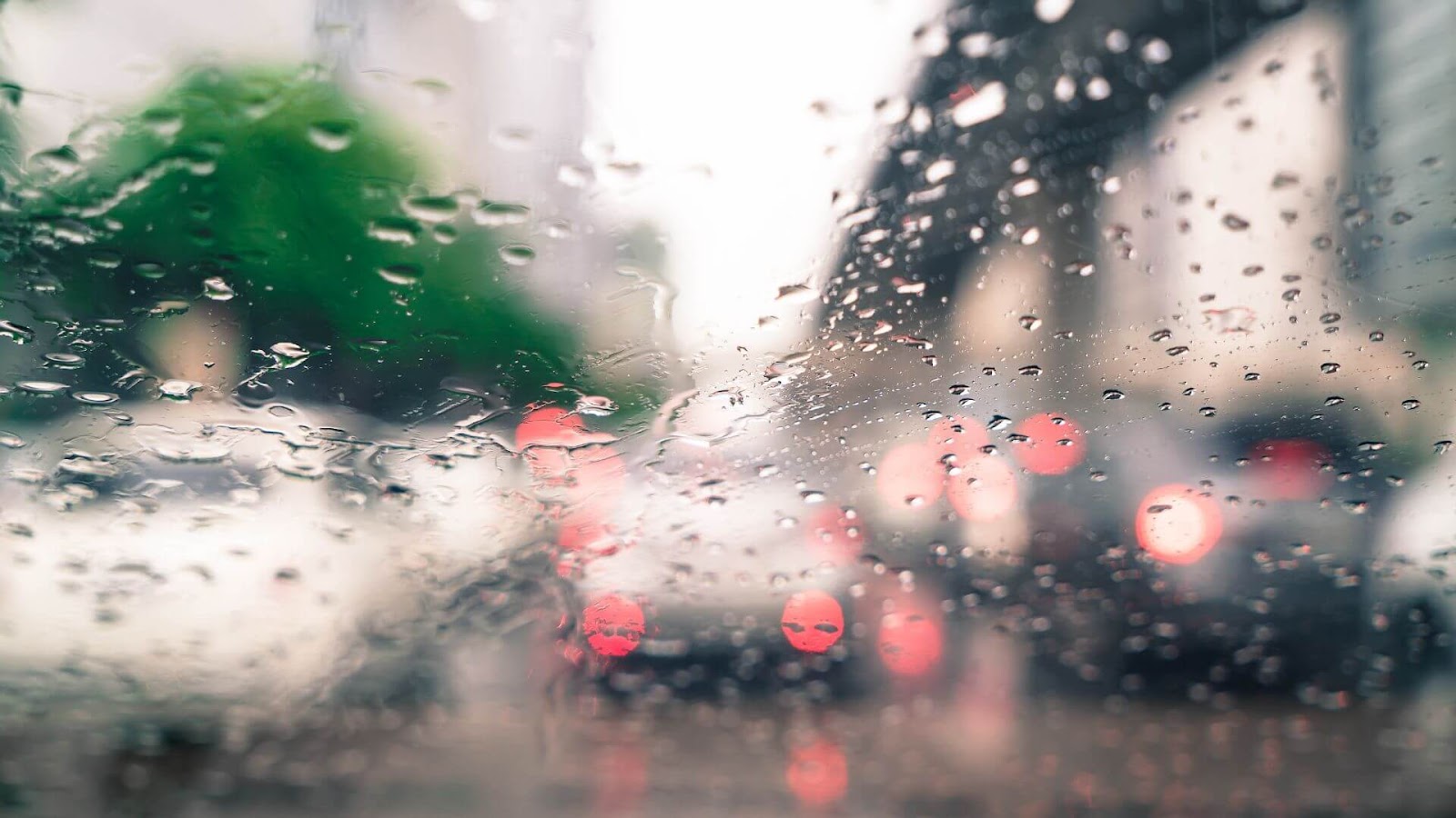 A imagem mostra uma vista através de um para-brisa de carro com muitas gotas de chuva, que estão borradas, criando um efeito de desfoque. Ao fundo, é possível distinguir luzes de carros em trânsito, que aparecem como pontos de luz vermelhos e brancos difusos. Há também a sugestão de folhagem verde do lado esquerdo, igualmente embaçada pela chuva e pelo desfoque. O ambiente geral da foto sugere um dia chuvoso ou garoa, com pouca visibilidade devido à água no vidro.