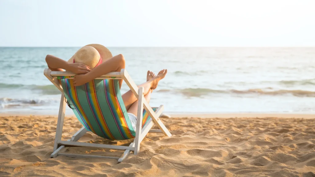Uma pessoa usando um chapéu de sol está relaxando em uma cadeira de praia listrada, de frente para o mar. A pessoa está sentada na areia da praia, com os pés levantados na direção do mar. O sol está baixo no horizonte, criando uma luz suave sobre a água e a areia.
