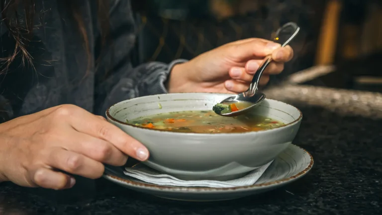 Uma pessoa segura uma tigela de sopa em um balcão ou mesa. Uma das mãos segura a borda da tigela, enquanto a outra usa uma colher para pegar um pouco da sopa, que contém vegetais.