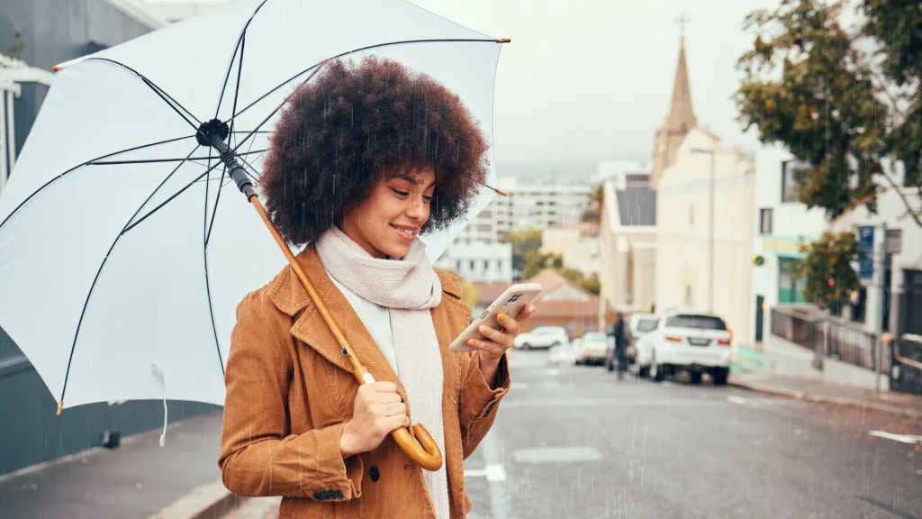 Uma mulher jovem com cabelo afro volumoso sorri enquanto olha para o celular sob uma chuva leve. Ela segura um guarda-chuva branco de cabo longo e veste um casaco marrom de veludo cotelê com um cachecol bege. Ao fundo, uma rua de cidade em declive com carros estacionados e prédios sob um céu nublado.