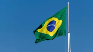 Bandeira brasileira ondulante com um losango verde e amarelo, com um globo azul e estrelas brancas, contra um céu azul claro.