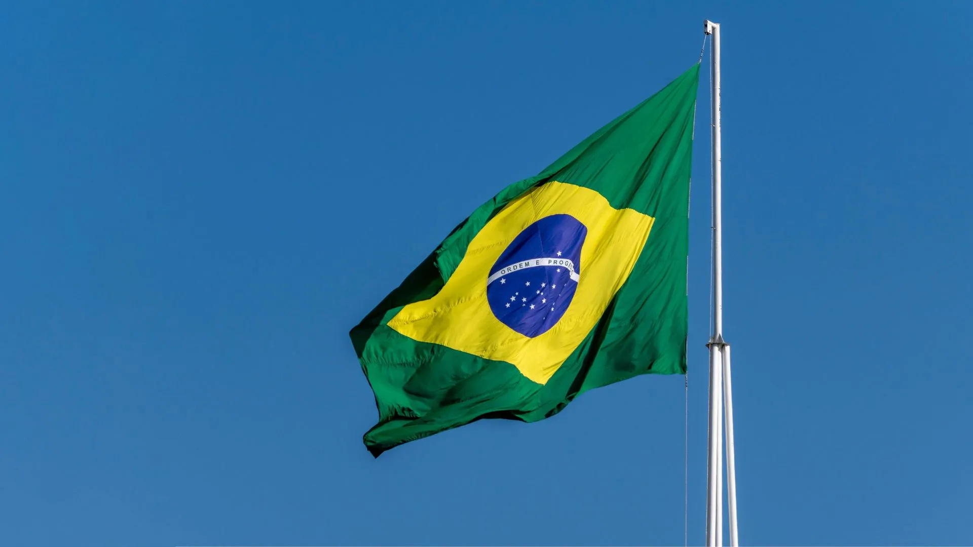 Bandeira brasileira ondulante com um losango verde e amarelo, com um globo azul e estrelas brancas, contra um céu azul claro.