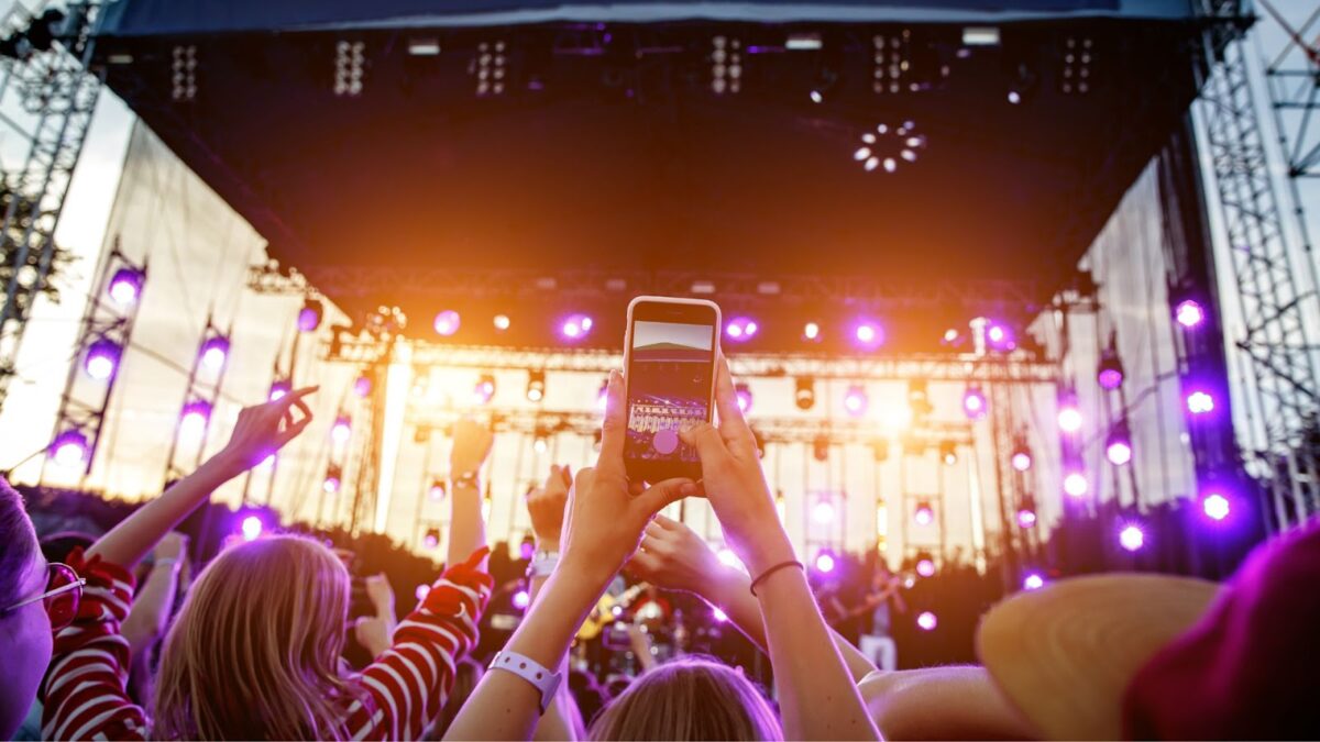 Pessoas em um show ou festival de música, com as mãos levantadas, algumas segurando smartphones para filmar o palco iluminado com luzes roxas e laranjas.