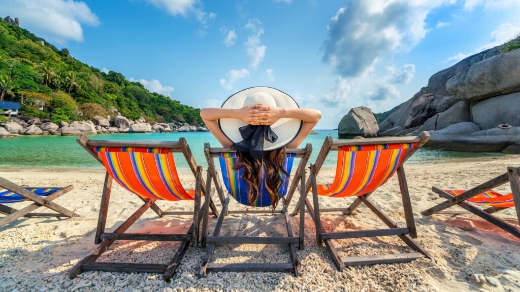 Uma pessoa vista de costas, usando um chapéu de sol branco, relaxa em uma cadeira de praia colorida, de frente para um mar de águas calmas e azul-turquesa, com uma encosta verde e rochas ao fundo.