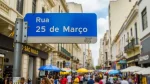 Placa de rua azul com a inscrição "Rua 25 de Março" em letras brancas, com a palavra "Rua" acima. Ao fundo, uma rua de São Paulo movimentada e cheia de pessoas. Há muitas barracas com guarda-sóis coloridos e lojas dos dois lados da rua.