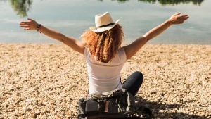 Uma mulher de chapéu de palha e cabelo ruivo e cacheado, sentada de costas para a câmera, de frente para um lago. Ela está com os braços abertos, em um gesto que sugere liberdade ou felicidade. Ela está sentada na margem de um lago, em uma área coberta por pequenas pedras e areia. A mulher usa uma blusa branca e calça escura, e há uma mochila escura na frente dela.