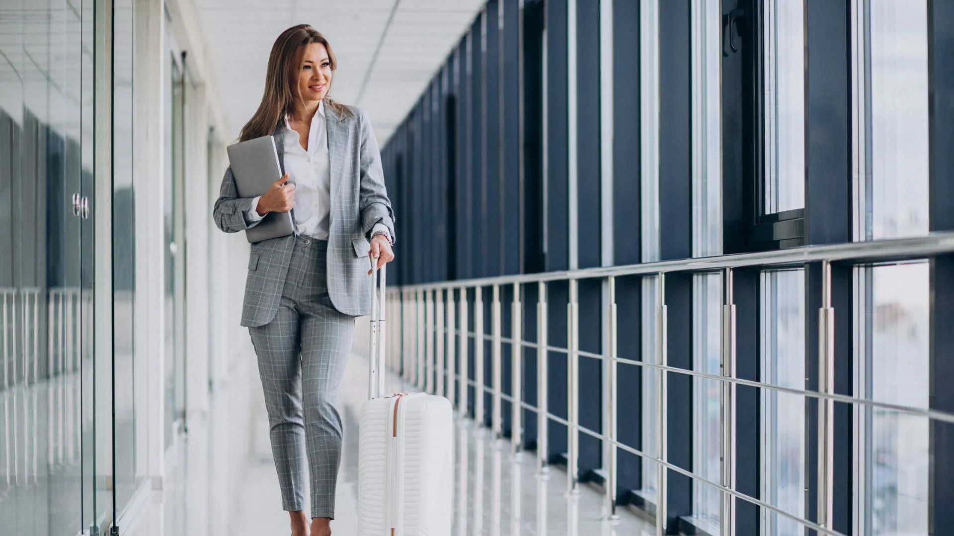 Mulher sorridente em traje de negócios cinza, com um laptop na mão, puxa uma mala de rodinhas branca por um corredor iluminado de aeroporto.